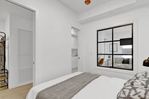 A bedroom with a large bed and a window with a view of a living room.