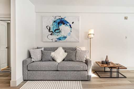 A grey couch with a white pillow and a blue painting on the wall.