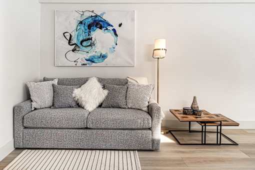 A grey couch with a white pillow and a blue painting on the wall.
