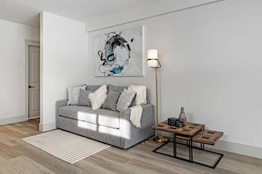 A grey couch with pillows and a blanket sits in a room with a painting on the wall and a small table with a vase and books.