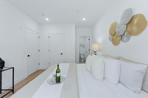 A white bedroom with a bed, a table with a bottle of wine and a tray, and a wall art piece.