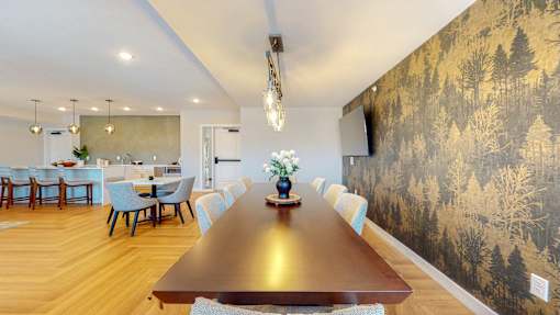 A long wooden table is surrounded by chairs in a room with a forest wallpaper.