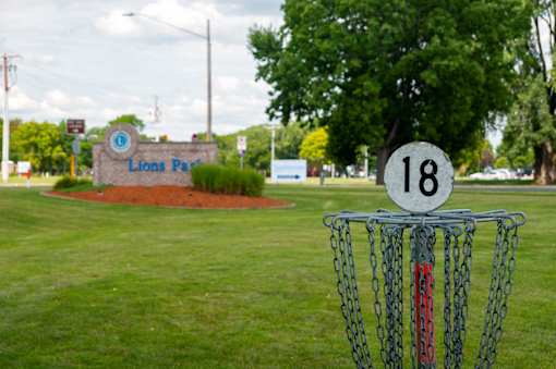 A disc golf basket with the number 18 on it is in the foreground of a grassy area with a sign that says Lions Park in the background.
