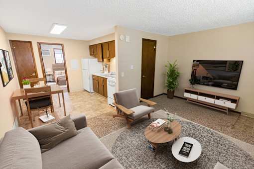 A living room with a grey couch, a coffee table, a dining table, a refrigerator, a television, and a potted plant.