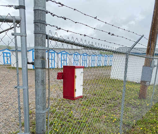 A red mailbox is attached to a chain link fence with barbed wire on top.