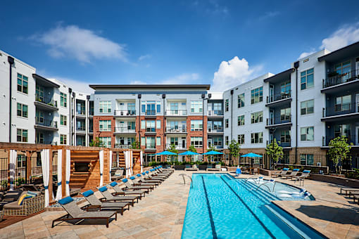 A large swimming pool surrounded by sun loungers and umbrellas in front of apartment buildings.
