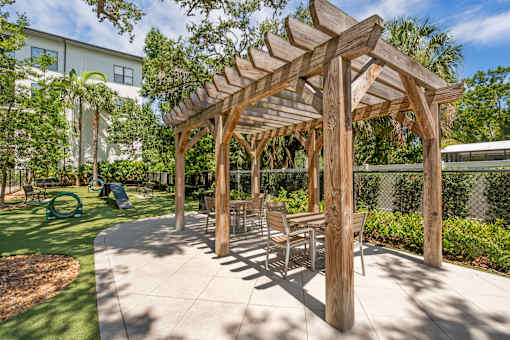 A wooden pergola with chairs and a table is in the middle of a sunny courtyard.