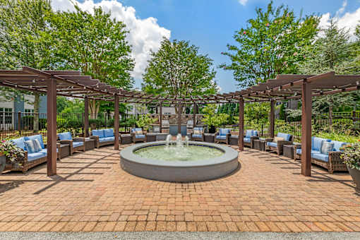 A fountain in the middle of a brick patio surrounded by trees and chairs.