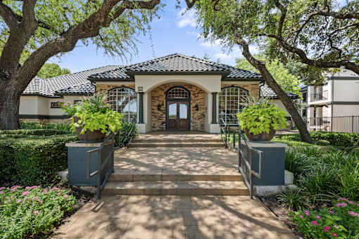 A house with a stone facade and a tiled roof is surrounded by greenery and has a pathway leading to the entrance.