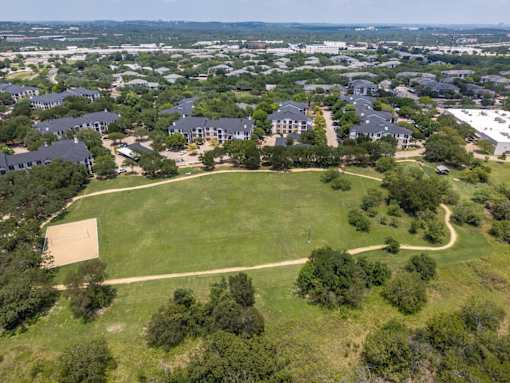 A large grassy field is surrounded by a winding path and buildings.