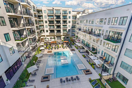 A large swimming pool is surrounded by lounge chairs and umbrellas in a courtyard surrounded by apartment buildings.