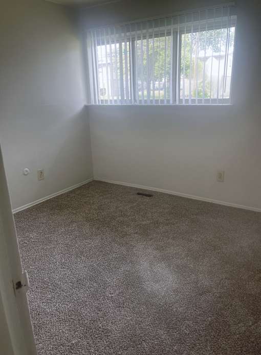 The bedroom in a 1 bedroom unit with a large window and gray carpet.