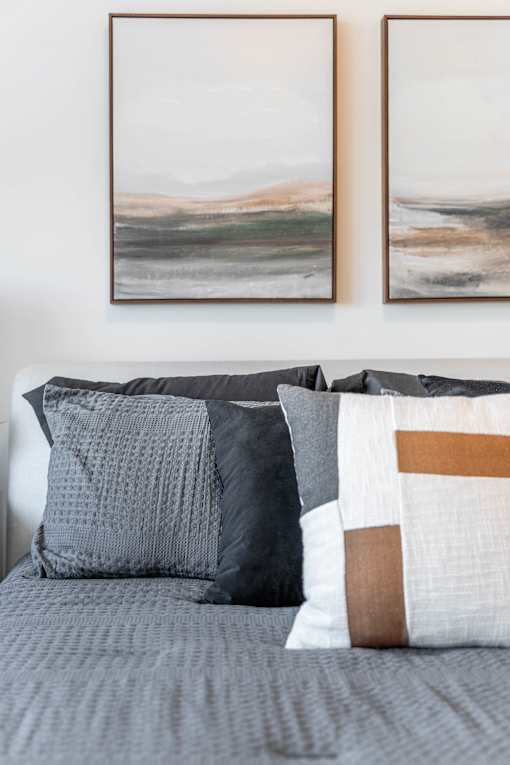 A bed with grey and white pillows and two landscape paintings on the wall.at Vibe Apartments at Arbour Lake, Calgary, T3G 4J2  