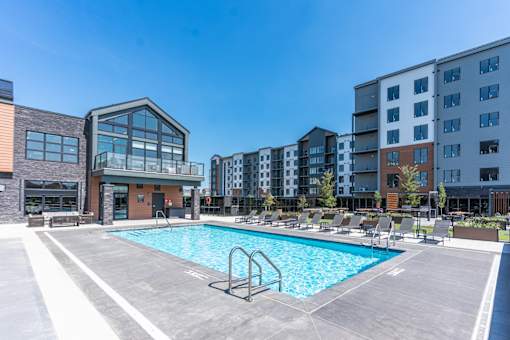 A swimming pool in a sunny day surrounded by chairs and buildings.