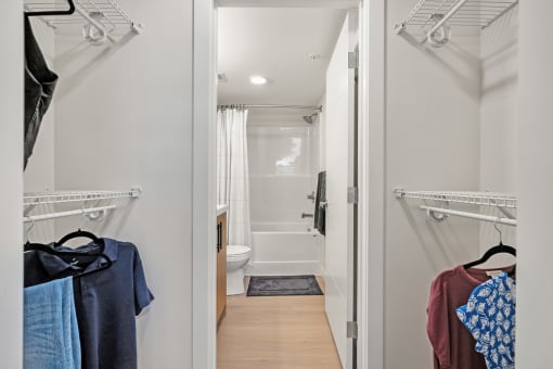 a walk in closet with clothes hanging on the wall and a bathroom in the background