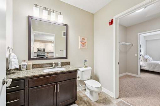A bathroom with a sink, mirror, toilet and a bedroom in the background.
