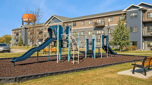 A playground with a blue slide and a brown bench in front of a building.