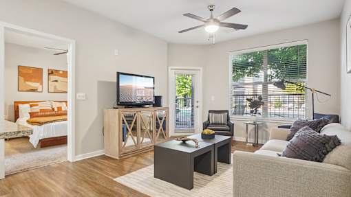 A living room with a grey couch, a wooden cabinet, a television, and a window with blinds.