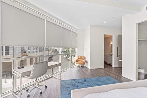 A room with a bed, desk, and chair with a view of the street outside.