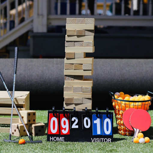 A game of Jenga is set up on a lawn with a scoreboard and ping pong paddles and balls nearby.