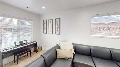 A black leather couch sits in a living room with a piano and two framed pictures on the wall.