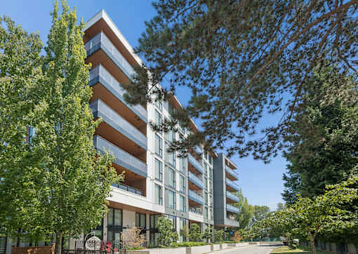 A modern multi-story building with balconies is surrounded by trees and greenery.