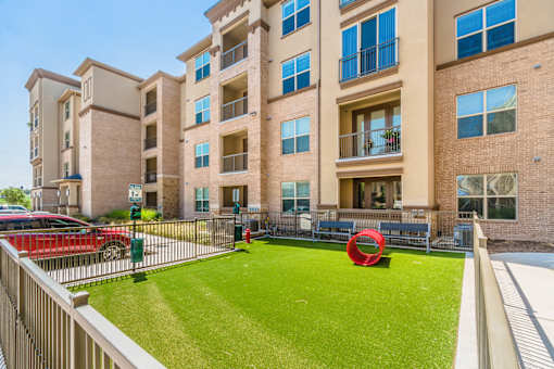A red car is parked in front of a building with a green lawn and a red sculpture at Atlas Point at Prestonwood Apartments , Carrollton, TX