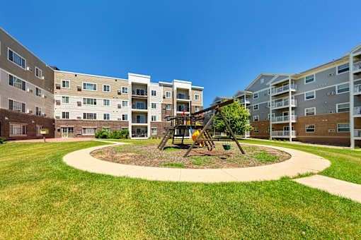 A playground with a swing set is in the middle of a grassy area in front of apartment buildings.