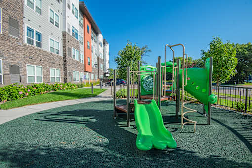 A playground with a green slide and a brown bench in front of a building.