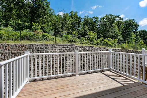 A white railing on a wooden deck with a stone wall and trees in the background.