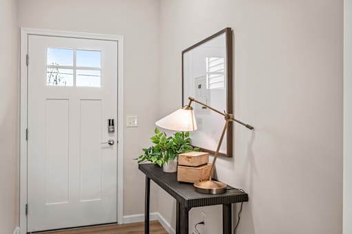 A white door is on the left of a table with a lamp and a plant on it.