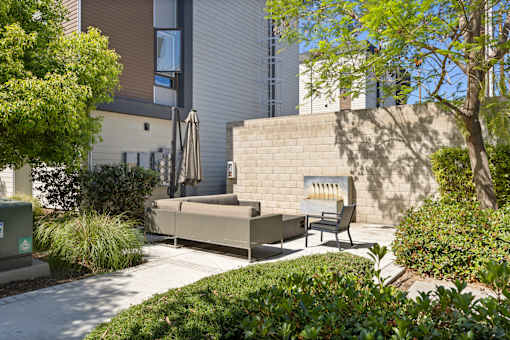 A patio with a couch and chairs is surrounded by green bushes.