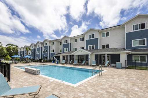 A swimming pool is surrounded by a patio and chairs in front of apartment buildings.