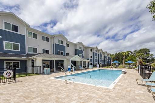 A swimming pool is surrounded by a patio and chairs in front of apartment buildings.