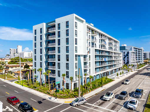 A modern white apartment building is situated on a street corner with cars parked and driving by.