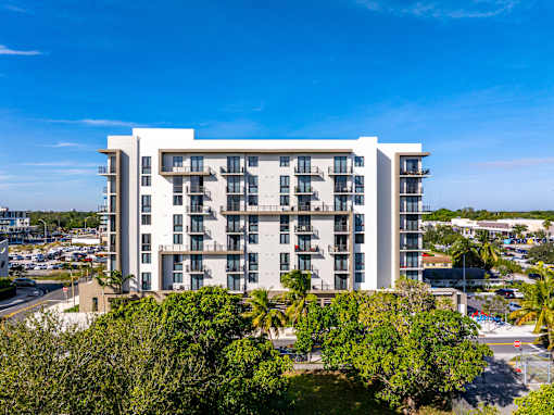 A large white building with balconies is surrounded by trees and a parking lot.