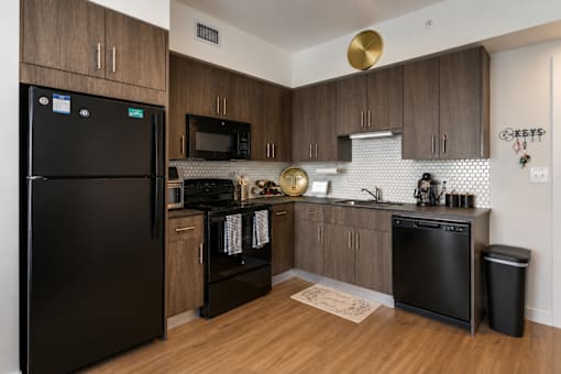 A black fridge sits in a kitchen with wooden cabinets and a black dishwasher.