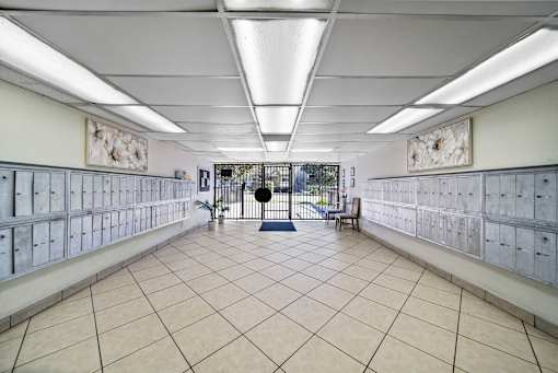 A room with a white tile floor and walls, with a row of mailboxes on the walls and a bench in the middle.