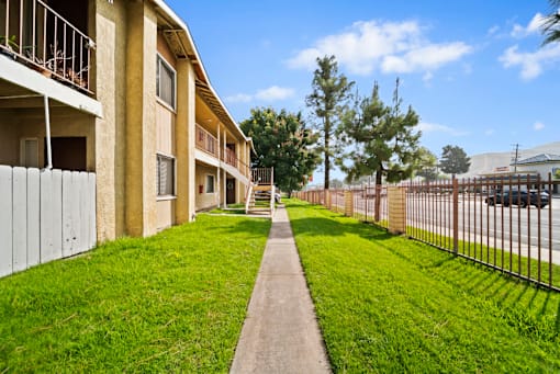 A long concrete walkway leads to a building with a metal fence on one side and a grassy area on the other.