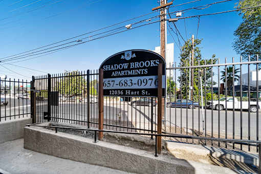 A sign for Shadowbrooks Apartments is displayed in front of a fence.