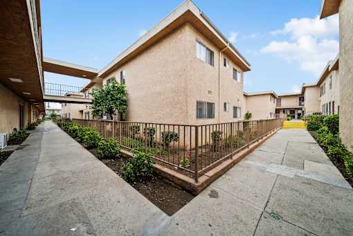 A concrete pathway leads to a building with a metal fence and a small tree in front.