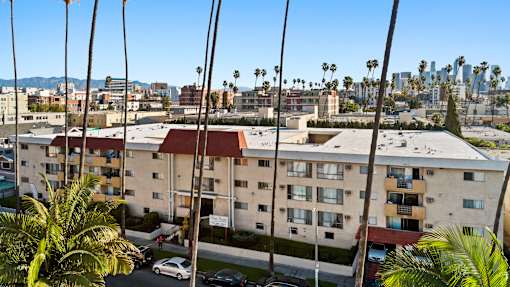 A view of a residential building with cars parked in front and palm trees in the foreground.