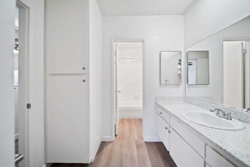 A white bathroom with a sink, mirror, and wooden floor.