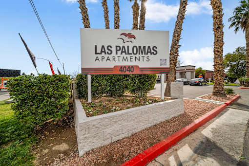 A sign for Las Palomas Apartments stands in front of a row of palm trees.