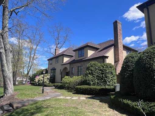 A house with a brown roof and a chimney is surrounded by green bushes and trees.
