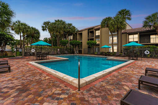 A pool surrounded by a brick patio and palm trees at Boca Colony Apartments, Boca Raton, FL