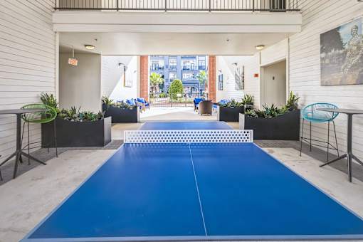 A blue ping pong table is in the middle of a room with a white wall and a painting hanging on i  at The Atlantic Memorial, Houston.