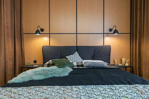 A bed with a floral comforter and a green pillow is in the foreground of a room with two wall lamps.