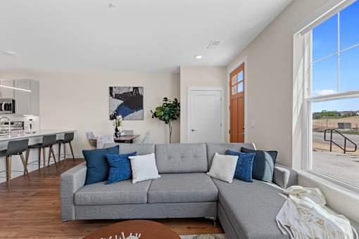 A grey couch with blue and white pillows sits in a living room with a kitchen area in the background at Tivoli Green Apartments & Townhomes, Mount Pleasant, Wisconsin, 53406