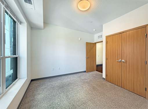 A room with a carpeted floor, two wooden doors, and a window with a view of a building outside.